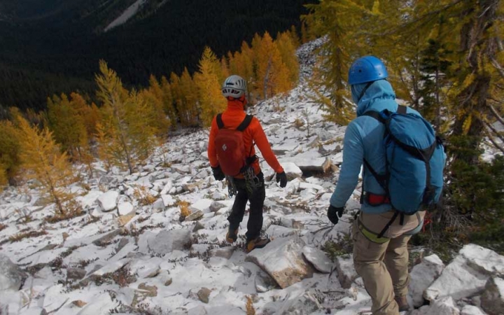 Two people wearing backpacks and helmets descend a rocky landscape dotted with orange trees